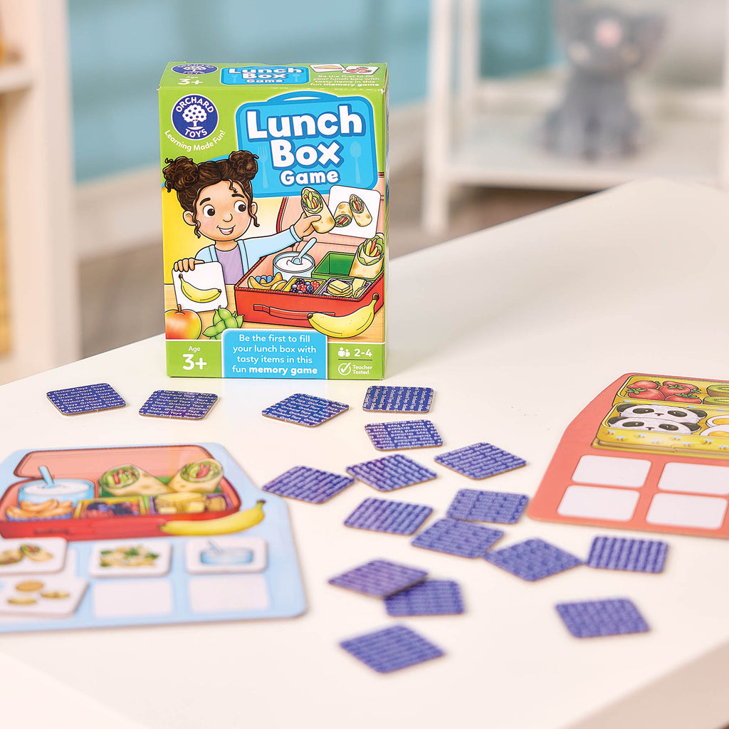 Organic Toys Lunch Box Game set displayed on a white table, showing the colorful game box, scattered blue square tokens, and two play cards in a bright indoor setting.