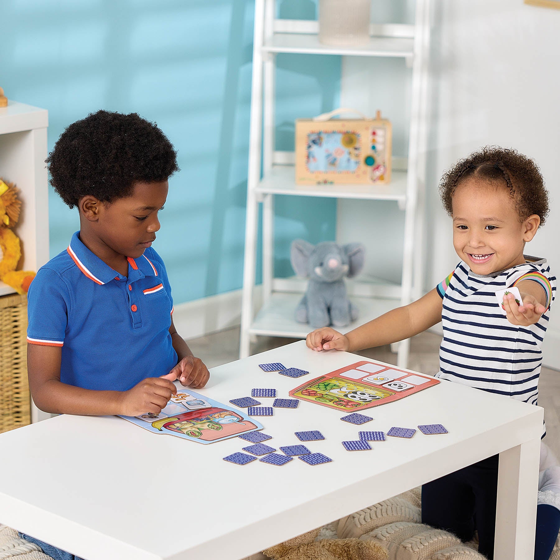 Two children playing with a colorful educational card game at a white table in a bright, modern playroom. One child is wearing a blue polo shirt and the other is wearing a navy and white striped shirt.