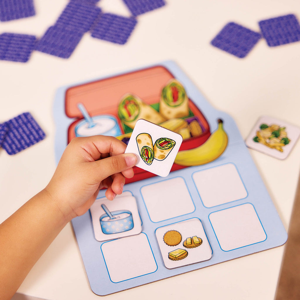 A child's hand holds a food identification card over a matching board game set on a white table. The game board shows a lunchbox with snacks and empty slots for matching cards.