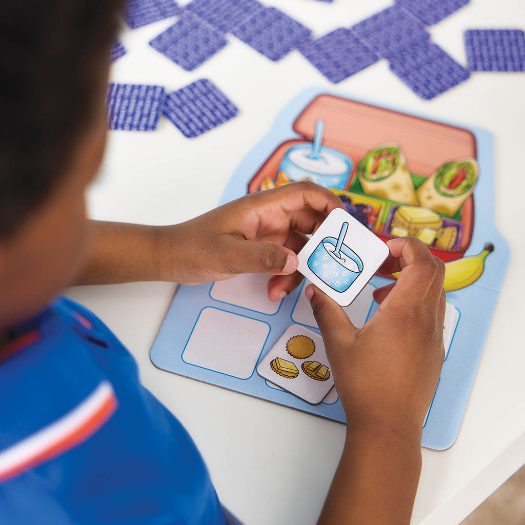 A child's hands holding a small illustrated food card over a colorful educational meal-planning board, with more matching cards scattered on a white table.