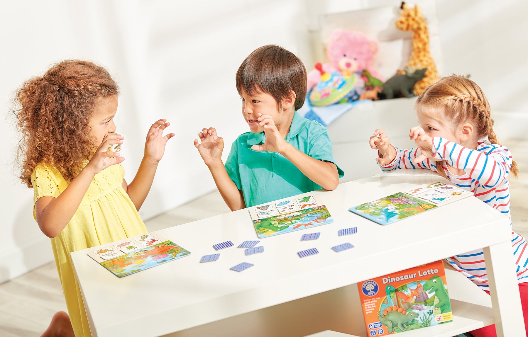 Three children playing with a colorful dinosaur card game set at a white table in a bright room. The box on the table reads 'Dinosaur Lotto'.