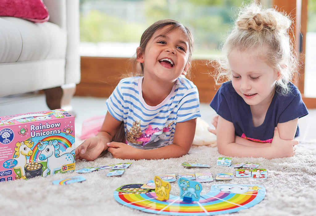 Two children playing with a board game on the floor, with a Rainbow Unicorns box in the background.