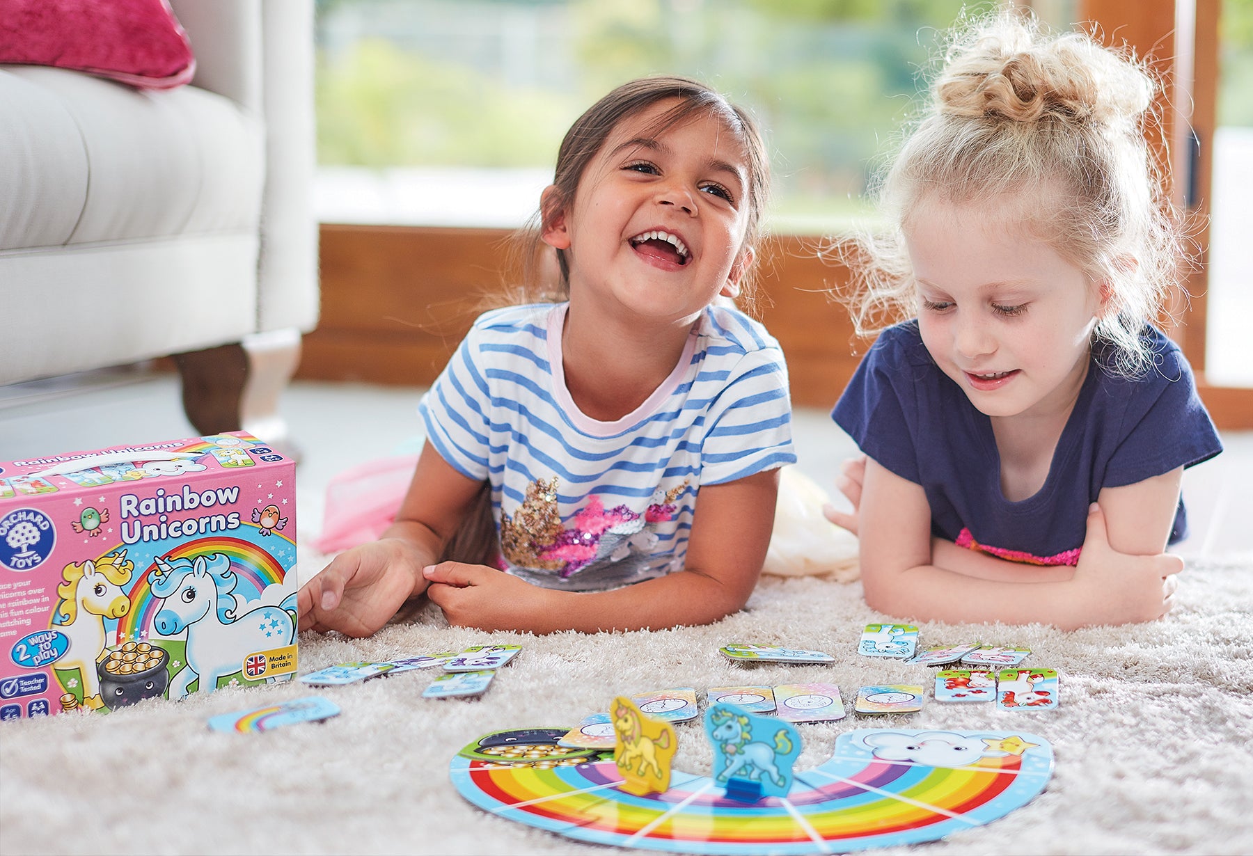 Two children playing with a board game on the floor, with a Rainbow Unicorns box in the background.