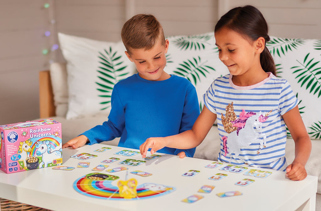 Two children playing with a board game on a table, with a box labeled 'Rainbow Unicorns' in the background.
