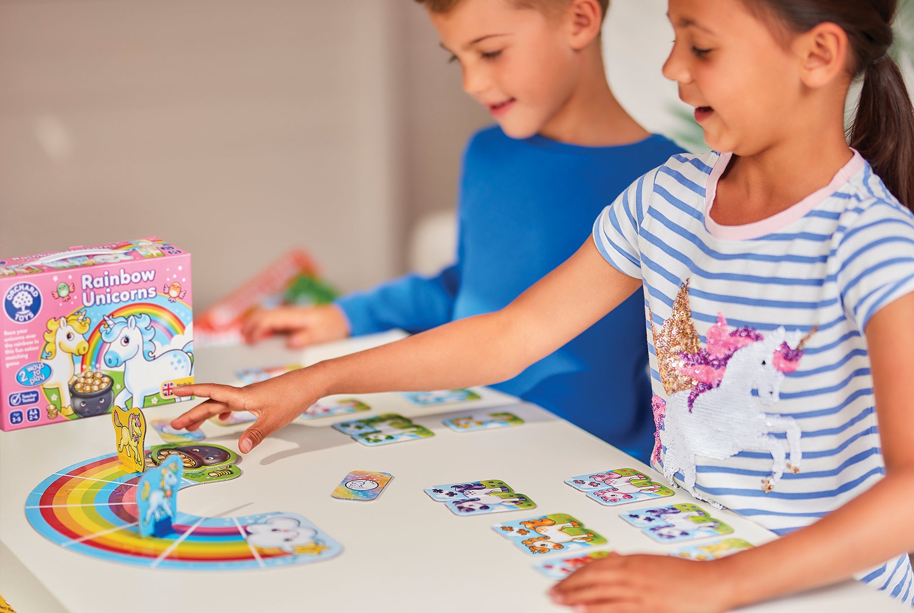 Two children playing with a board game featuring rainbow unicorns on a table.