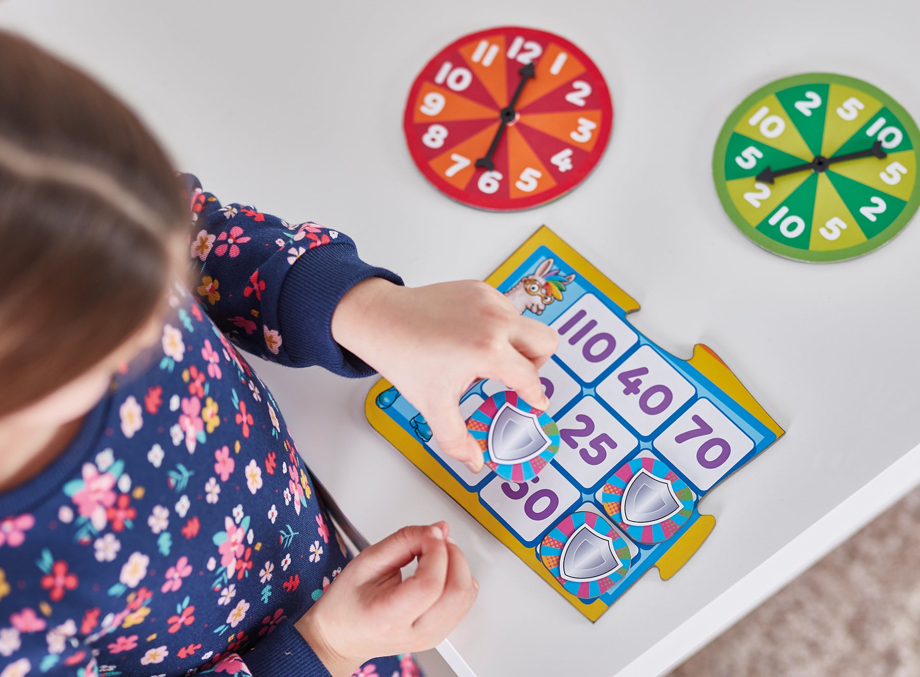 Child interacting with educational number board game on a white surface with colorful clocks in the background.
