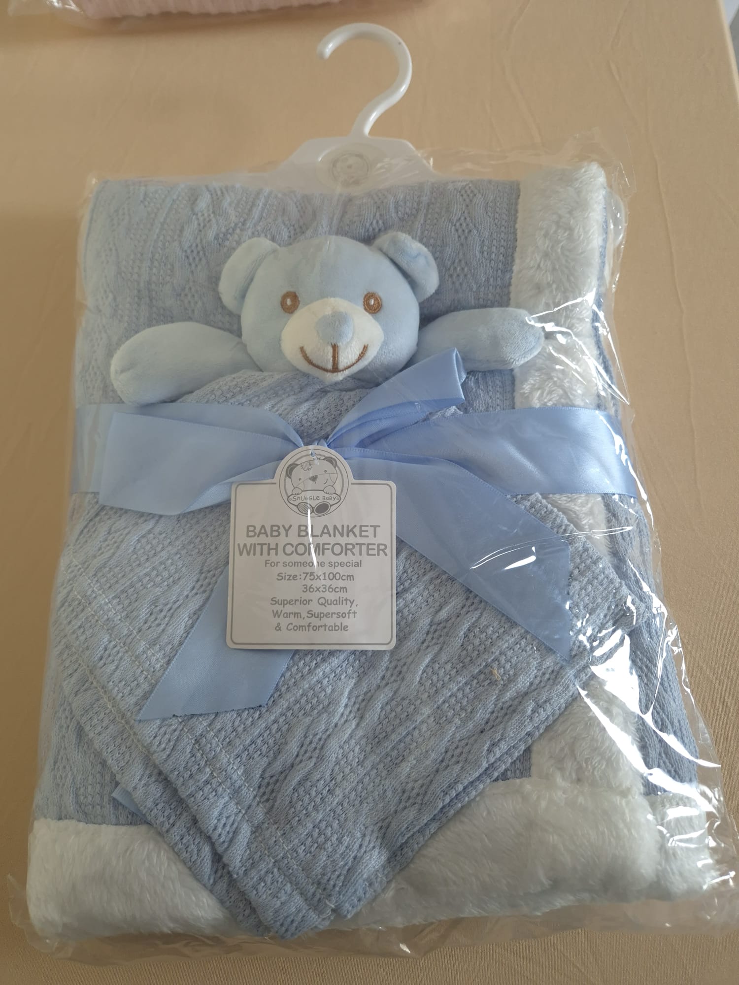 A light blue and white knitted baby blanket with a matching plush teddy bear attached, wrapped in clear plastic packaging with a white hanger and ribbon bow. A product label is attached in the center.