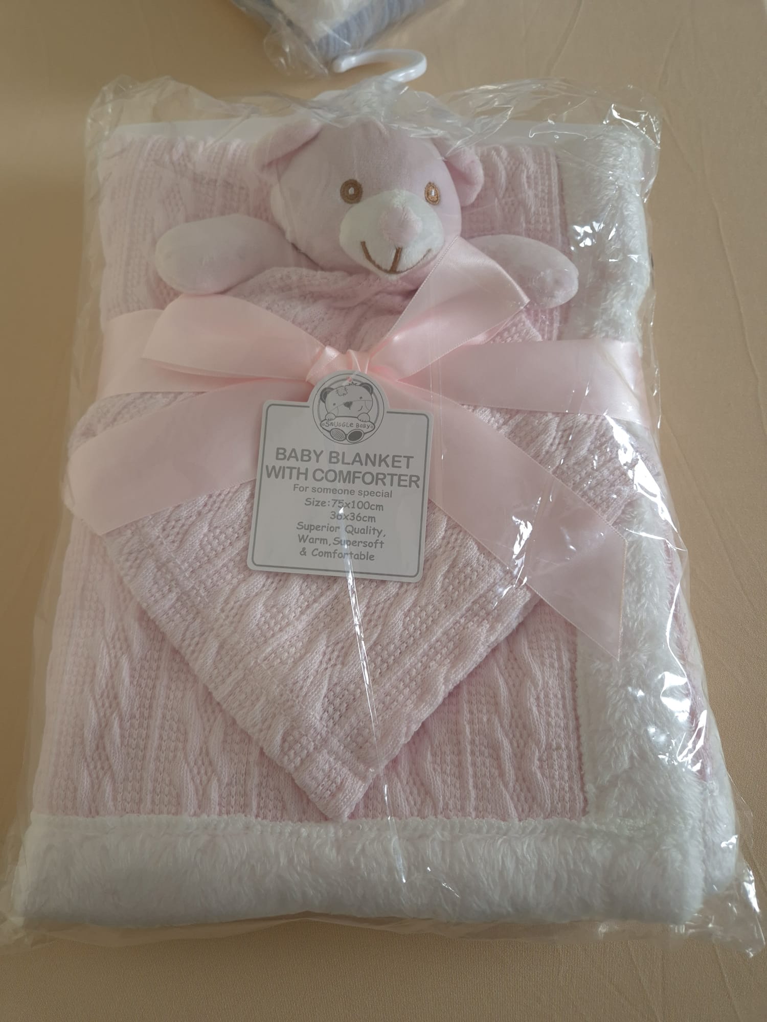 A packaged baby blanket set with a pink knitted blanket, matching plush bear comforter, and a pale pink ribbon bow, wrapped in clear plastic with a product tag attached.